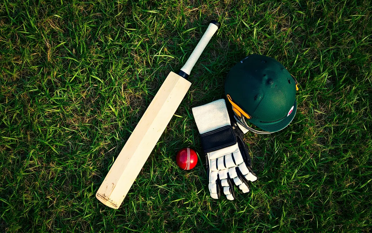 Overhead flat-lay of a cricket batting setup on green grass: bat, gloves, helmet and one red ball arranged in order