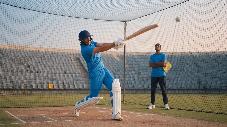 Batter in a plain blue training kit practicing a lofted straight drive into a net while a coach watches from the side