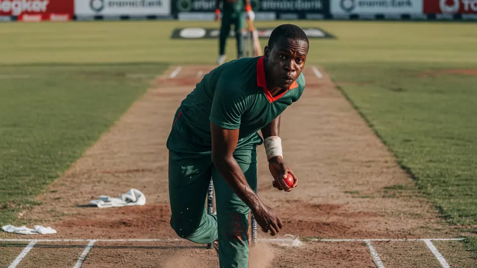 Close-up of a bowler in a green kit releasing a red ball in his delivery stride, dust lifting off the pitch with a batter blurred at the far crease